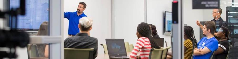 A room of students watch as an instructor points at a screen