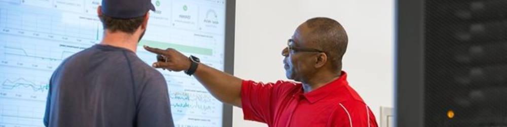 A man points at a large screen displaying charts and graphs as another man looks on.